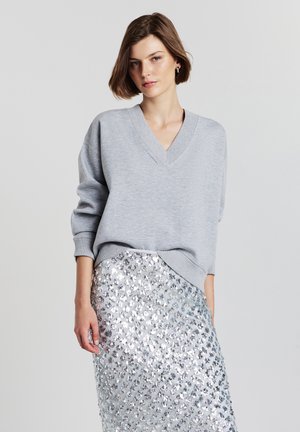 Donna con i capelli corti castani, che indossa un maglione grigio chiaro a V e una gonna di paillettes argentate, in piedi contro uno sfondo chiaro e semplice.