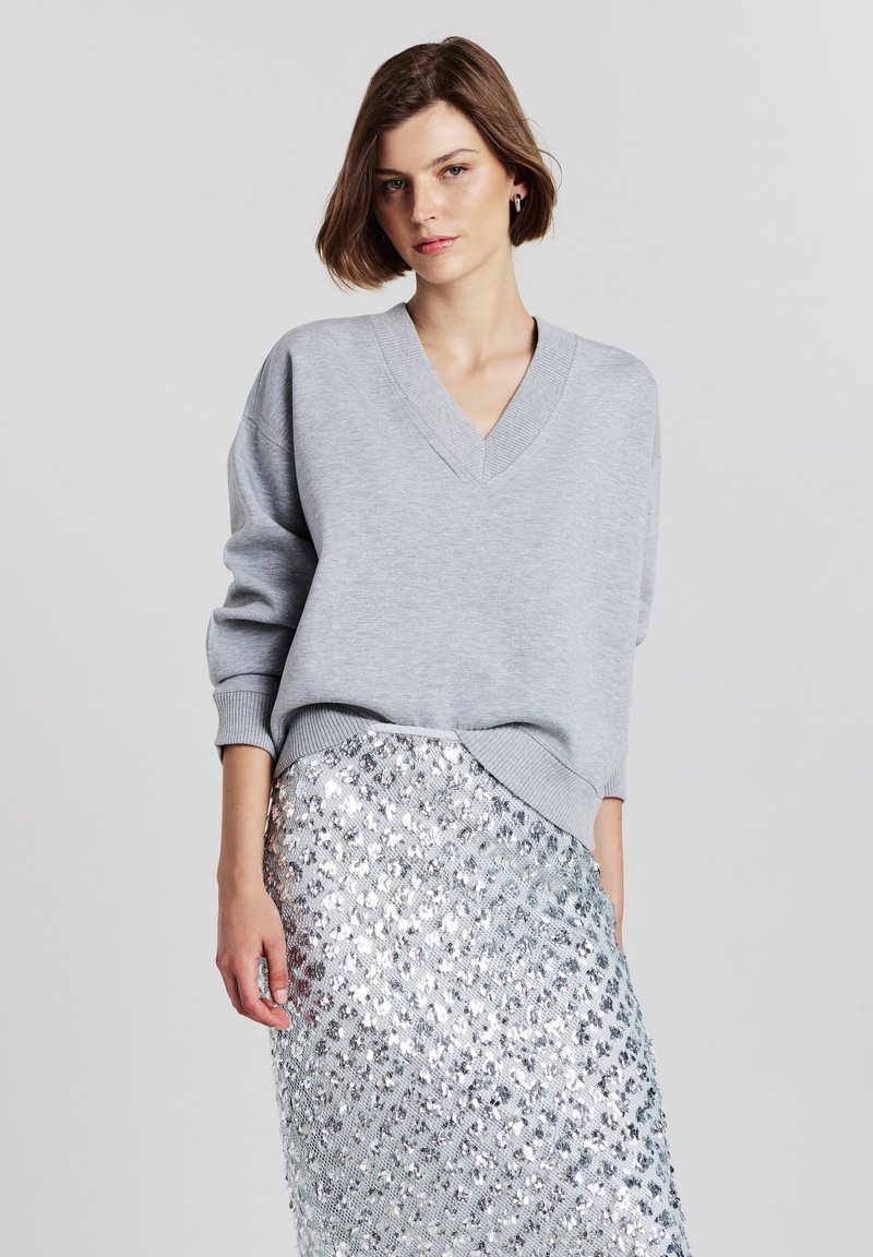 Donna con i capelli corti castani, che indossa un maglione grigio chiaro a V e una gonna di paillettes argentate, in piedi contro uno sfondo chiaro e semplice.