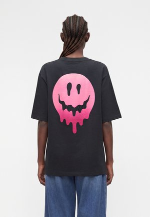 Άτομο με πλεξούδες που φοράει μαύρο μπλουζάκι με ένα μεγάλο ροζ μουτράκι που στάζει στην πλάτη, σε συνδυασμό με μπλε τζιν.