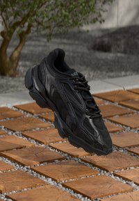 Black athletic shoe with textured leather upper, featuring streamlined design, fabric lining, and rubber sole. Floating above a stone path.