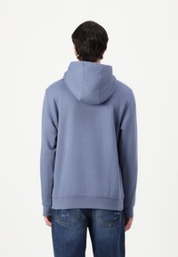 Felpa in cotone blu con cappuccio regolabile, polsini e orlo a coste, e una texture morbida. Indossata su jeans in denim blu.