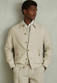 Chaqueta de lino beige con cuello, botones de madera, bolsillos laterales y cintura elástica, combinada con un suéter de punto ligero.