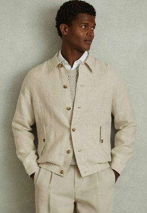 Beige linnen jas met een kraag, houten knopen, zijzakken en een elastische tailleband, gedragen over een licht gebreid truitje.
