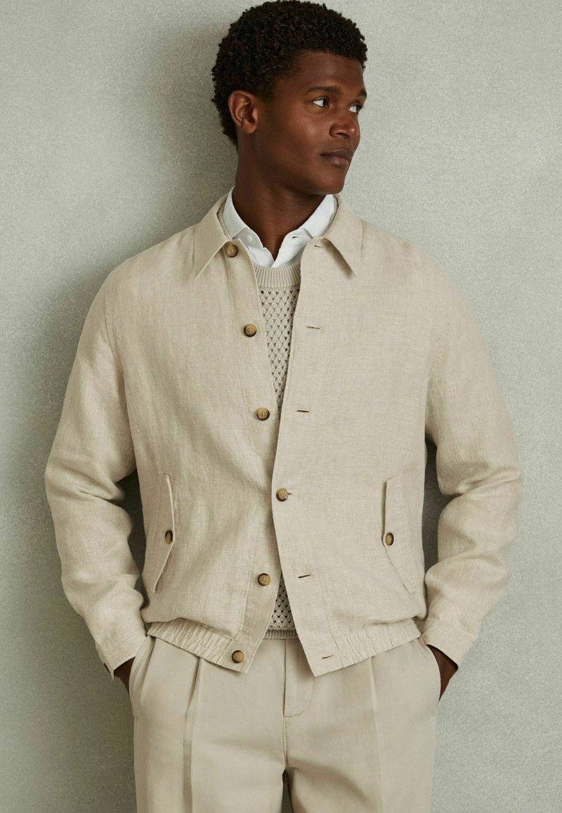 Chaqueta de lino beige con cuello, botones de madera, bolsillos laterales y cintura elástica, combinada con un suéter de punto ligero.