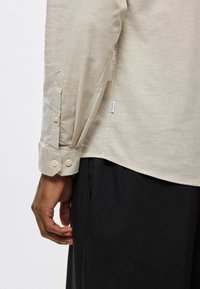 Πουκάμισο μακρυμάνικο από ανοιχτό μπεζ ύφασμα. Διαθέτει μύτες στα μανίκι α με δύο κουμπιά και μια μικρή ετικέτα στη ραφή πλευράς.