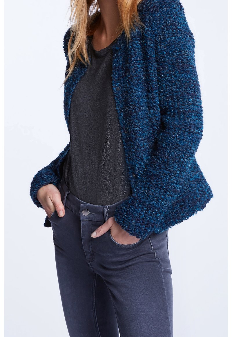 Personne portant un cardigan bleu texturé, un haut noir et un jean gris foncé avec les mains dans les poches, visible des épaules aux hanches.