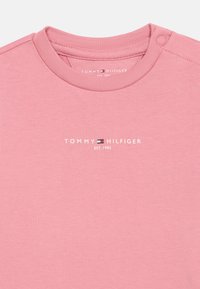 Helles rosa Baumwoll-T-Shirt mit Rundhalsausschnitt und Druckknopfdetails. Auffällige weiße "TOMMY HILFIGER"-Markenbeschriftung mit kleinem Logo darunter.