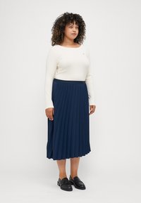 Marinblå plisserad kjol med en slät midja, tillsammans med en krämfärgad långärmad topp. Modellen bär svarta loafers och står mot en neutral bakgrund.