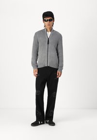 Cardigan de cremallera acanalado gris combinado con pantalones sueltos negros con gráficos bordados en contraste. Modelo lleva gafas de sol negras.
