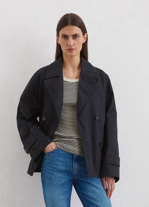 Veste double boutonnage bleu marine foncé avec grands revers, accents de boutons et manches retroussées, portée sur une chemise rayée et un jean bleu.