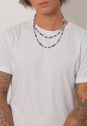 Colliers à perles superposées avec de petites perles multicolores et un accent de perle blanche centrale, portés sur un t-shirt blanc uni.