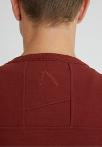 Pull bordeaux à encolure ronde, en tricot texturé, avec une couture diagonale subtile et une broderie rouge contrastante au niveau de l'encolure arrière.