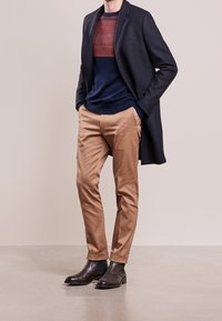 Marineblauer Wollmantel über einem marinen und kastanienbraun gestreiften Pullover, kombiniert mit hellbraunen, taillierten Hosen und dunkelbraunen Leder-Ankle-Boots.