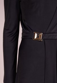 Robe noire à manches longues avec une finition satinée et une ceinture à boucle carrée en or, mettant en valeur une texture douce et un design élégant.