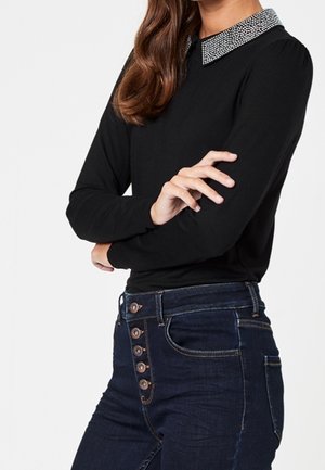 Femme portant un haut à manches longues noir avec un col en strass et un jean taille haute bleu foncé avec des boutons apparents.