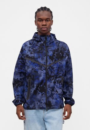 Ung mand iført en blå og sort pixeleret camouflagesjakke med hætte, lynet op, og lyseblå jeans, stående foran en ensfarvet baggrund.