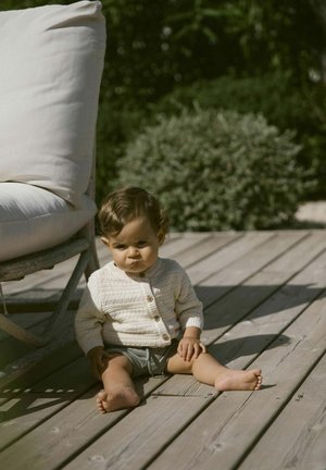 Strick-Cardigan in Creme mit Holzknöpfen, kombiniert mit grauen Shorts. Baby sitzt auf einer Holzterrasse, neben einem großen Kissen.