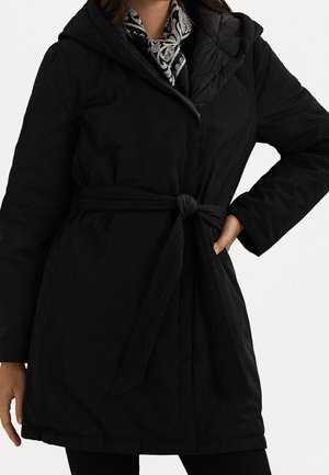 Femme portant un manteau à capuche noir ceinturé sur une écharpe à motif cachemire, une main sur la hanche et l'autre détendue.