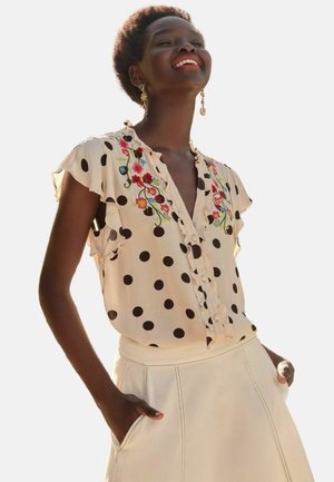 Beige blouse met zwarte stippen, bloemige borduursels, gerimpelde mouwen en een knopenlijst aan de voorkant. Gecombineerd met een lichtgekleurde rok.