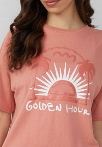 T-shirt en coton rose avec un graphique représentant un soleil blanc et un palmier, accompagné du texte "HEURE D'OR" en dessous. Design classique à col rond.