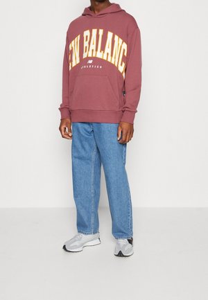 Henkilö, jolla on päällään viininpunainen New Balancen hupullinen paita, väljät siniset farkut ja harmaat sekä valkoiset lenkkarit, seisoo yksinkertaisen valkoisen taustan edessä.