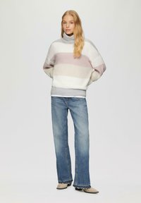 Oversized coltrui van zachte brei met horizontale pastelkleurige strepen; gecombineerd met lichtblauwe flared jeans en beige schoenen.