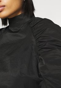 Femme portant un haut noir à col montant avec des manches bouffantes, montrant les lèvres, la ligne de la mâchoire et les cheveux courts et foncés.