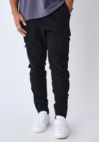Pantalones cargo negros con múltiples bolsillos, hechos de una tela ligera, diseño ajustado y combinados con zapatillas blancas.