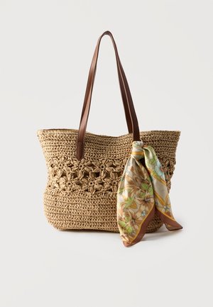 Sac cabas beige tissé avec des poignées en cuir marron et un foulard en soie à motif floral noué à une poignée, posé sur un fond uni.
