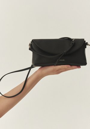 Mână care ține o geantă crossbody neagră din piele, cu clapetă și curea reglabilă, pe un fundal simplu, deschis.