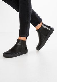 Botas de tobillo de cuero negro con un diseño sin cordones, paneles laterales elásticos y una suela de goma negra gruesa. Combinadas con jeans oscuros sobre una superficie blanca.