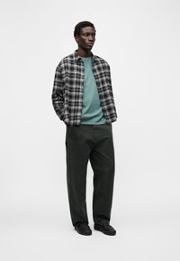 Άνδρας που στέκεται με τα χέρια στις τσέπες φορώντας μαύρο και άσπρο πουκάμισο με καρό, τιρκουάζ μπλούζα, άνετο σκούρο παντελόνι και μαύρα παπούτσια σε απλό φόντο.