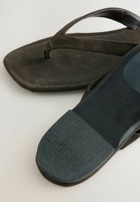 Tamsiai rudos šlepetės su tekstūruota padu ir plonomis dirželių detalėmis, padėtos ant šviesaus fono, demonstruojančios savo dizainą ir medžiagas.