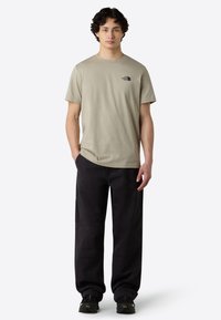 T-shirt di cotone beige chiaro con maniche corte, caratterizzata da un piccolo logo nero sul petto; abbinata a pantaloni neri larghi.