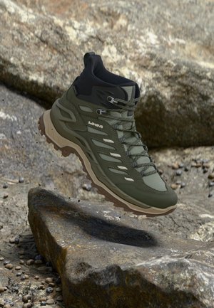 Olivgrüner Wanderstiefel mit schwarzer Knöchelstütze und beiger Sohle, der über felsigem Gelände schwebt.
