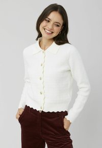 Cardigan blanc à côtes avec une bordure et un col ondulés, orné de boutons dorés sur le devant. Le mannequin porte un pantalon en velours côtelé bordeaux.