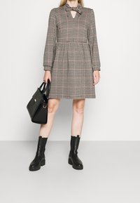 Robe à carreaux dans des teintes beige, noir et rouge. Présente un col haut, un détail en forme de trou de serrure, des manches longues et une jupe évasée. Associée à un sac à main noir et des bottes.