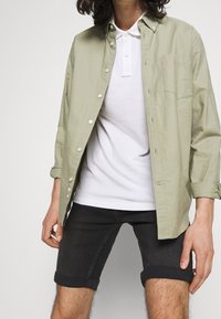 Chemise boutonnée vert clair sur un polo blanc, accompagnée de shorts en denim foncé avec des manchons retroussés et une poche avant sur la chemise.