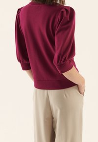 Bordeauxfarbener Pullover mit Puffärmeln, weichem Textil und einer taillierten Taille, kombiniert mit hellbeigen Hosen, die Hände in den Taschen.