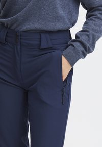 Pantalons de sport navy extensibles avec une texture lisse, dotés d'une poche latérale zippée, de fermetures à bouton et d'une coupe ajustée à la taille.