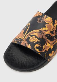 Chinelo preto com uma sola texturizada, preta e uma tira larga com um padrão floral ornado em dourado e laranja sobre um fundo escuro.