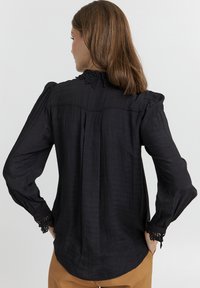 Blusa nera a maniche lunghe in tessuto testurizzato. Presenta un colletto bordato in pizzo e accenti in pizzo sui polsini, con pieghe sottili sul retro.