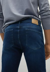 Dark blue denim jeans with a smooth texture, straight cut, and two back pockets. A tan leather label is visible at the waistband.