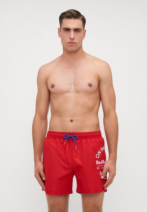 Red swim shorts with a textured finish, featuring a blue drawstring and white text graphics. Mid-thigh length and elastic waistband.