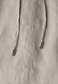 Tissu en lin beige avec deux cordons lâches se terminant par de petites agrafes métalliques cylindriques argentées, couture verticale centrée.