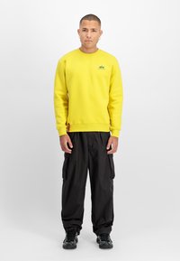 Gul genser med ribbede mansjetter og nederkant, med en liten logo. Kombinert med svarte cargobukser. Jevn tekstur og avslappet passform.