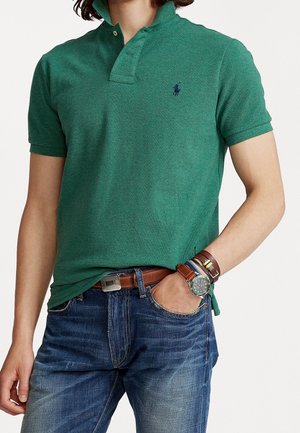 Grünes Poloshirt mit Kragen, kurzen Ärmeln und kleinem Logo. Kombiniert mit blauer Jeans und mehreren Armbändern am Handgelenk.