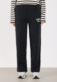 Czarne dresowe spodnie z elastycznym pasem, z białym napisem "TOKYO 1963", prosty krój i miękka tekstura. Połączone z szarymi butami sportowymi.