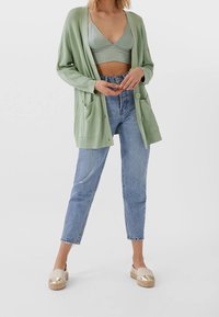 Cardigan verde chiaro con apertura frontale, due tasche e chiusura a bottone. Abbinato a jeans a vita alta blu chiaro e scarpe dai toni neutri.
