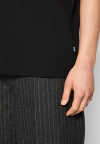 T-shirt en coton noir avec un ourlet arrondi, présentant une petite étiquette blanche "BOSS" sur le côté, associé à un pantalon gris à rayures.